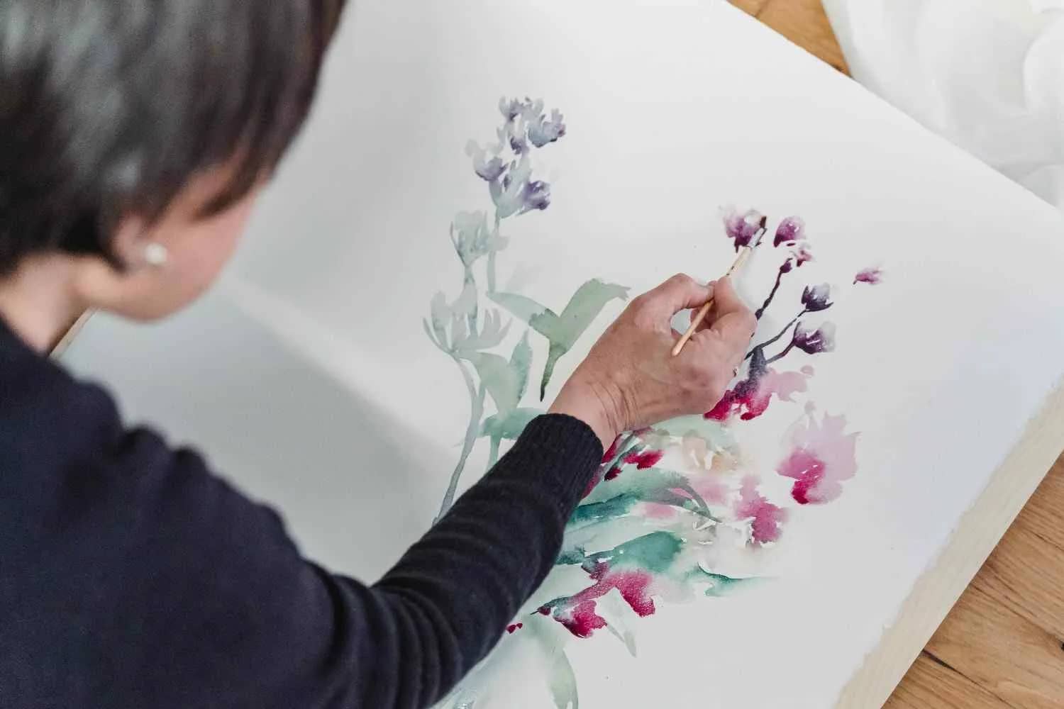 A woman's hands painting a floral watercolor scene, with a focused expression.
